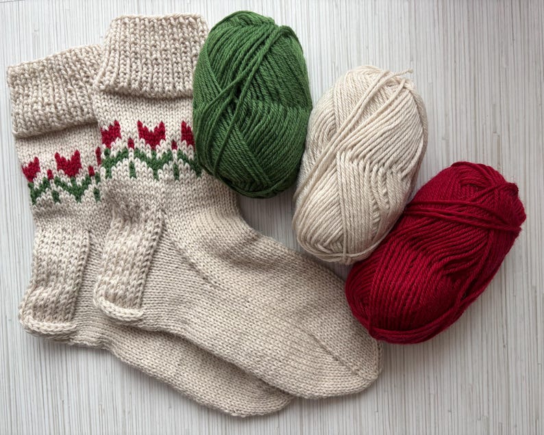 May include: A pair of beige knitted socks with a red and green floral pattern. The socks are lying on a white surface with three balls of yarn: green, beige, and red.