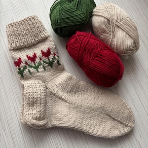 May include: A beige knitted sock with a green and red tulip pattern. Three balls of yarn are shown in the background: green, beige, and red.
