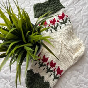 May include: A pair of knitted socks with a white background and a green and red tulip pattern. The socks are folded and lying on a white surface.