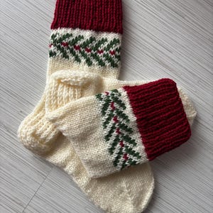 May include: A pair of hand-knitted Christmas stockings. The top cuff is a deep red, transitioning to a cream-colored body with a green and red fir tree pattern. The stockings are made of a thick, textured yarn.