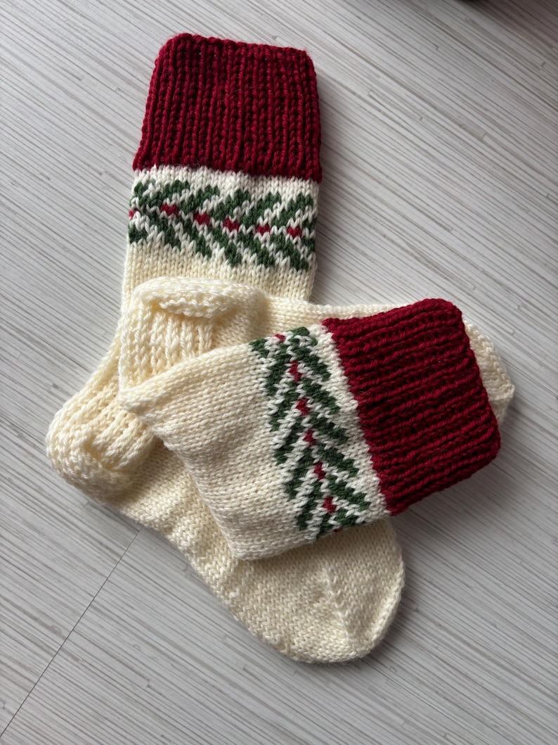 May include: A pair of hand-knitted Christmas stockings. The top cuff is a deep red, transitioning to a cream-colored body with a green and red fir tree pattern. The stockings are made of a thick, textured yarn.