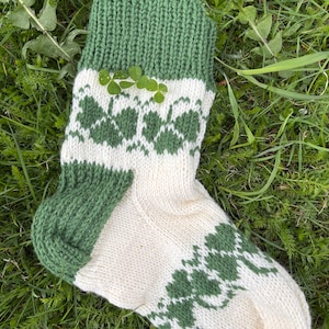 May include: A pair of knitted socks with a white background and green clover designs. The socks have a green cuff and a green heel.