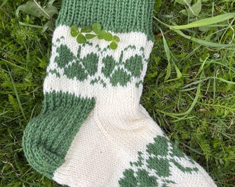 May include: A pair of knitted socks with a white background and green clover designs. The socks have a green cuff and a green heel.