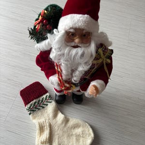 May include: A Santa Claus figurine with a red suit and hat, white beard, and glasses stands next to a cream-colored knitted Christmas stocking with a red cuff and green and red design. The Santa figure holds a sack.
