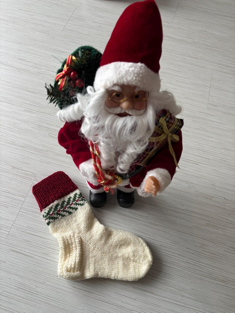 May include: A Santa Claus figurine with a red suit and hat, white beard, and glasses stands next to a cream-colored knitted Christmas stocking with a red cuff and green and red design. The Santa figure holds a sack.