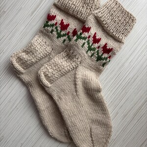 May include: A pair of beige knitted socks with a red and green floral pattern. The socks are folded together and have a ribbed cuff.
