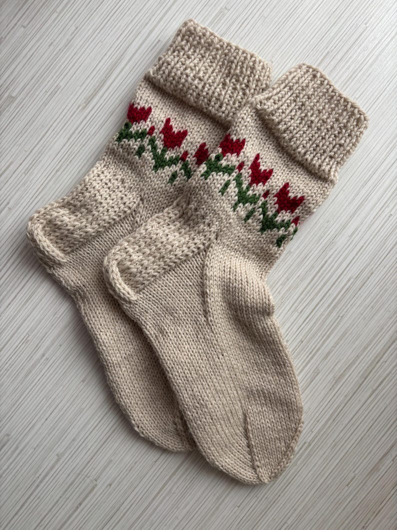 May include: A pair of beige knitted socks with a red and green floral pattern. The socks are folded together and have a ribbed cuff.