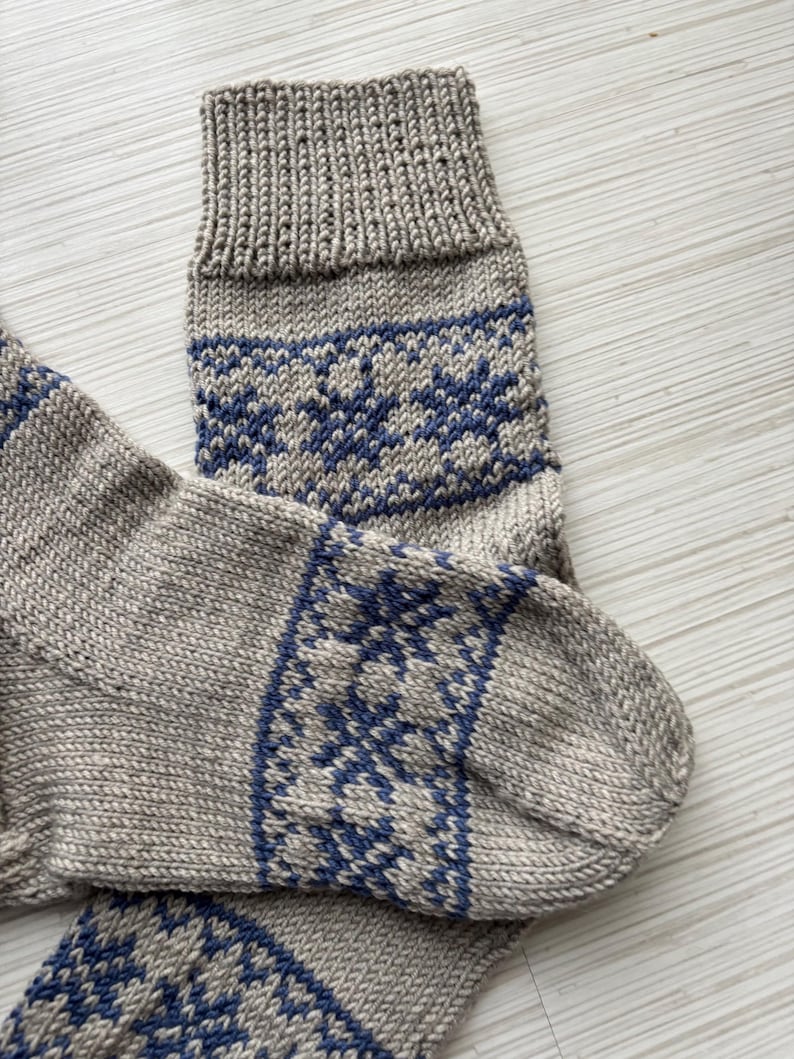 May include: A pair of knitted socks with a grey base and blue snowflake pattern. The socks are folded and the top of the sock is visible.