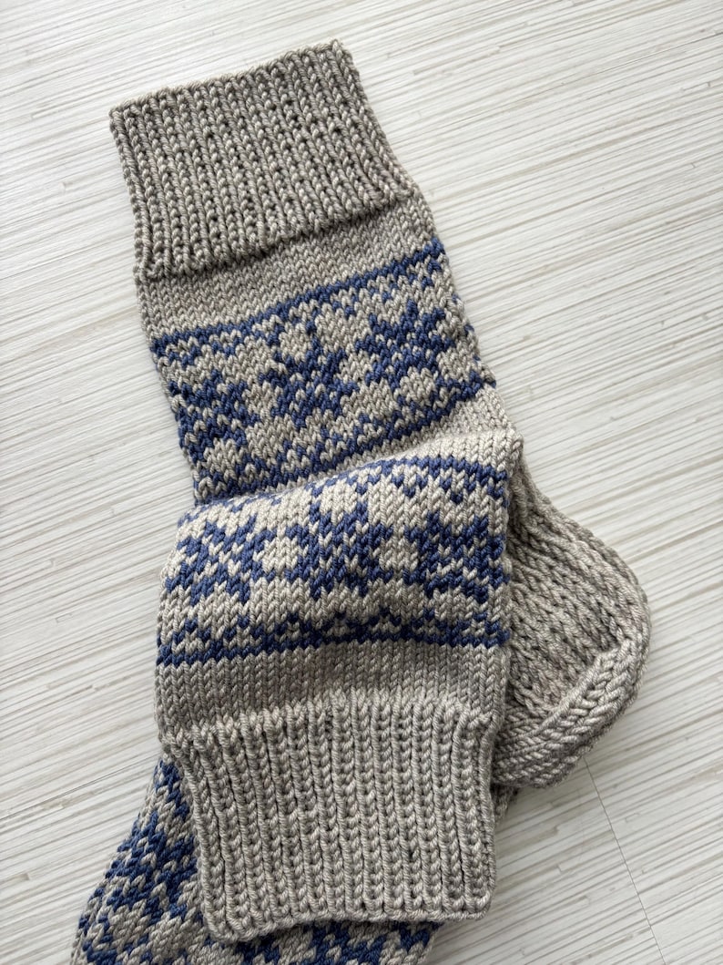 May include: A pair of knitted socks with a grey and blue geometric pattern. The socks are folded and the top of the sock is visible.