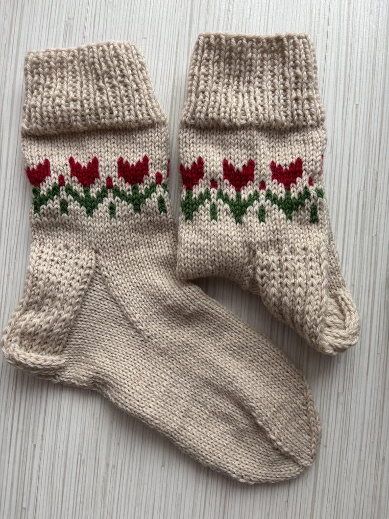 May include: A pair of beige knitted socks with a red and green floral pattern. The socks are made of a soft, comfortable material and have a ribbed cuff.