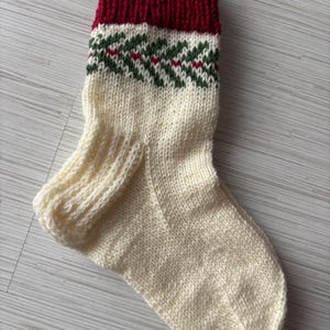 May include: A hand-knitted Christmas stocking in cream, with a deep red cuff. The stocking features a green and red geometric pattern near the cuff. The cozy, festive design is perfect for holiday decor.