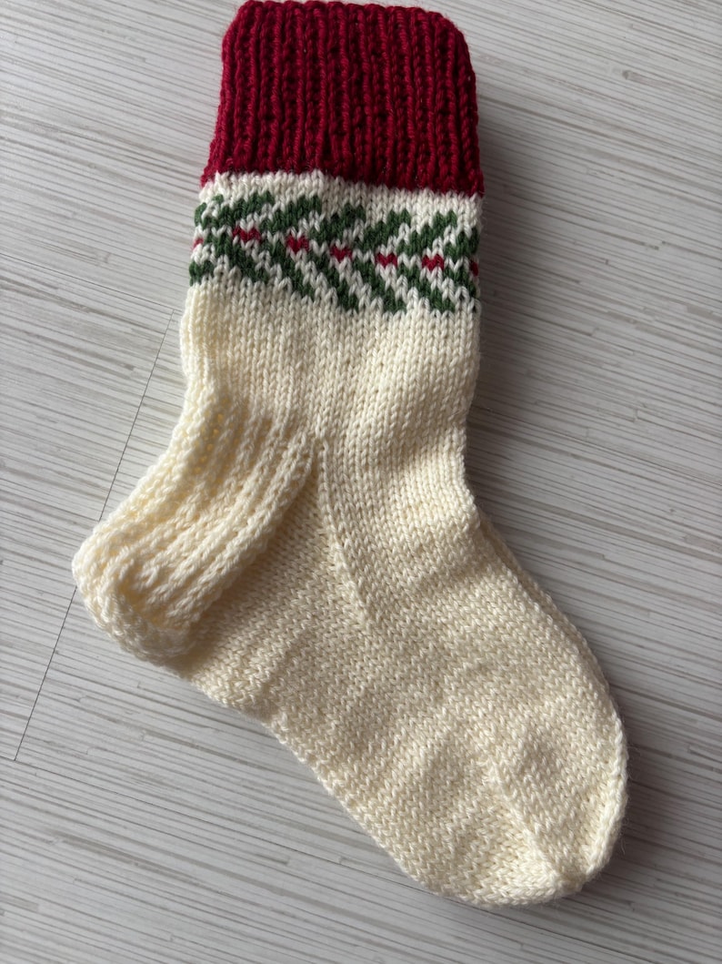 May include: A hand-knitted Christmas stocking in cream, with a deep red cuff. The stocking features a green and red geometric pattern near the cuff. The cozy, festive design is perfect for holiday decor.