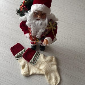 May include: A Santa Claus figurine with a red hat and coat stands above two cream-colored knitted Christmas stockings. The stockings have a red cuff with a green and white patterned design. The Santa Claus figurine is holding a bag.