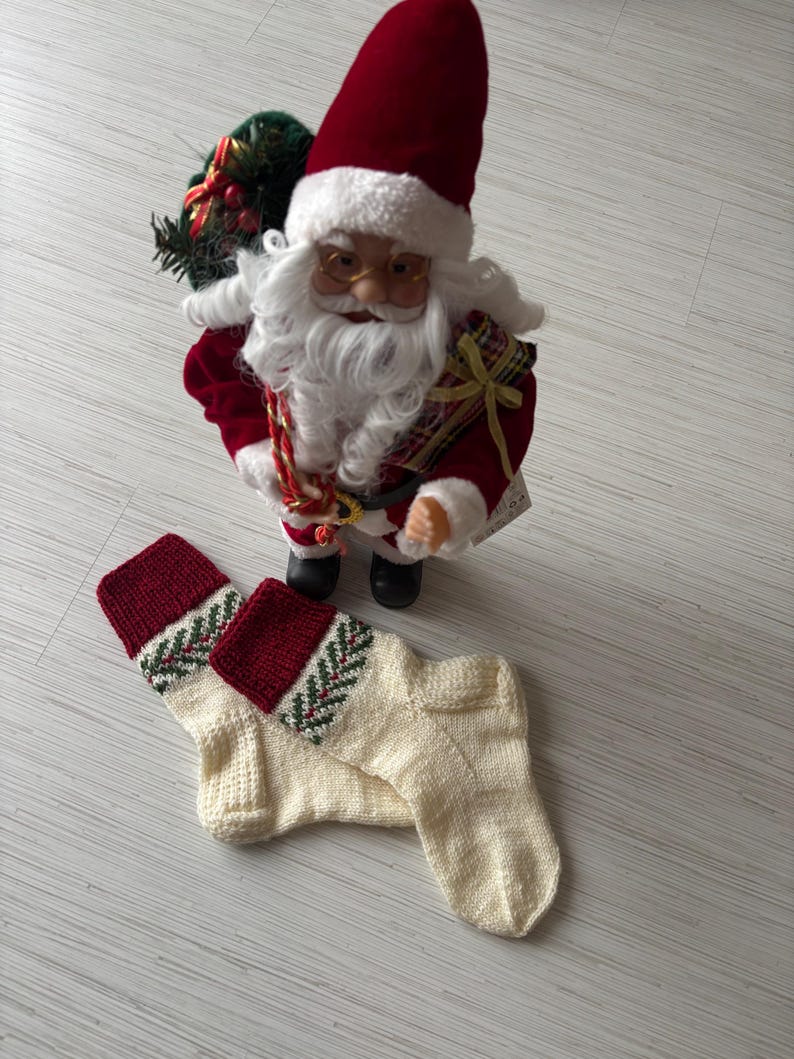 May include: A Santa Claus figurine with a red hat and coat stands above two cream-colored knitted Christmas stockings. The stockings have a red cuff with a green and white patterned design. The Santa Claus figurine is holding a bag.