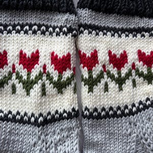 May include: A pair of knitted socks with a white background and a pattern of red tulips with green stems. The socks have black and gray stripes at the top and bottom.