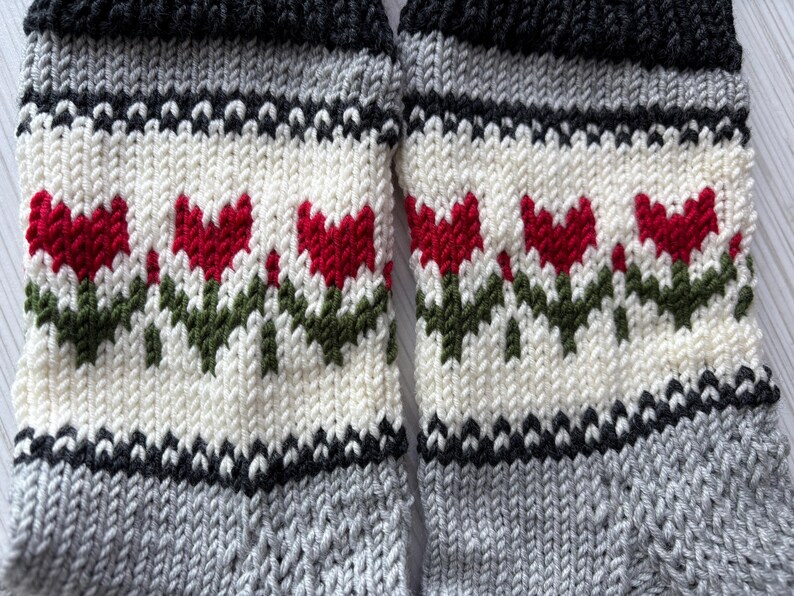 May include: A pair of knitted socks with a white background and a pattern of red tulips with green stems. The socks have black and gray stripes at the top and bottom.