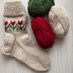 May include: A beige knitted sock with a red and green tulip pattern, along with three balls of yarn in beige, green, and red.