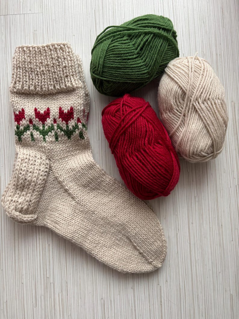 May include: A beige knitted sock with a red and green tulip pattern, along with three balls of yarn in beige, green, and red.