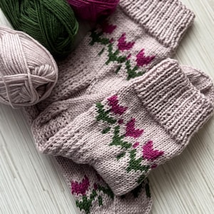 May include: A pair of light pink knitted socks with a floral design in magenta and green. The socks are folded, with three balls of yarn in green, magenta, and light pink in the background. The socks are handmade.