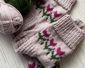 May include: A pair of light pink knitted socks with a floral design in magenta and green. The socks are folded, with three balls of yarn in green, magenta, and light pink in the background. The socks are handmade.