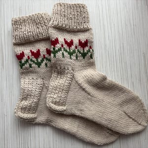 May include: A pair of beige knitted socks with a red and green floral pattern. The socks are made of a soft, comfortable material and have a classic design.