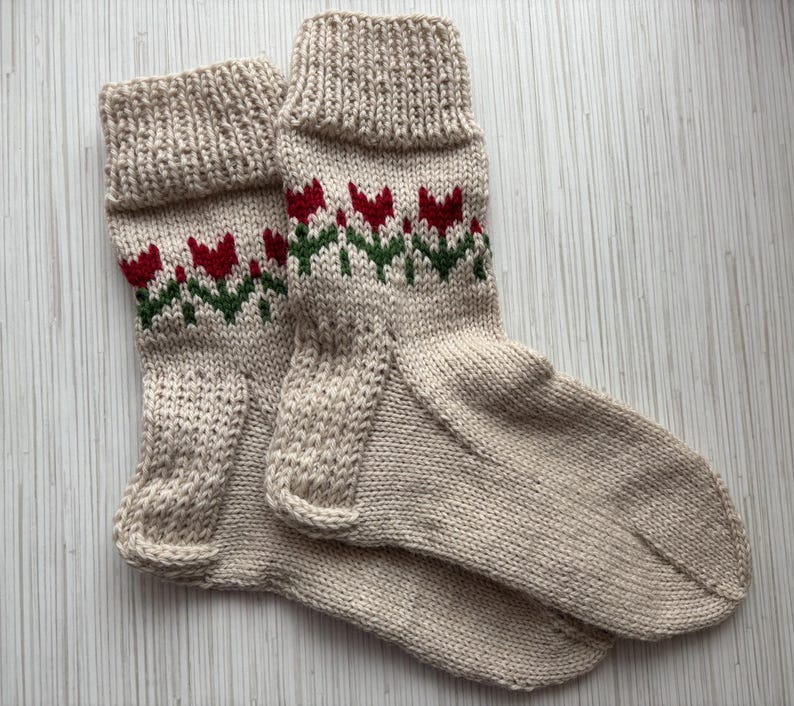 May include: A pair of beige knitted socks with a red and green floral pattern. The socks are made of a soft, comfortable material and have a classic design.