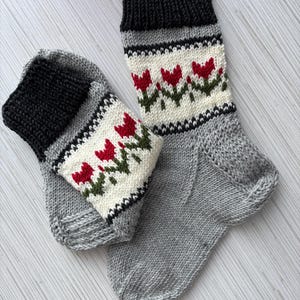 May include: A pair of hand-knitted socks with a grey base and a white band featuring red tulips. The socks have black cuffs.
