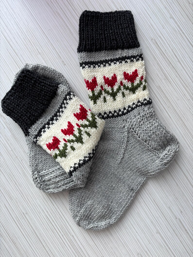 May include: A pair of hand-knitted socks with a grey base and a white band featuring red tulips. The socks have black cuffs.