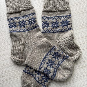 May include: A pair of grey knitted socks with a blue snowflake pattern. The socks are folded together and are lying on a white surface.