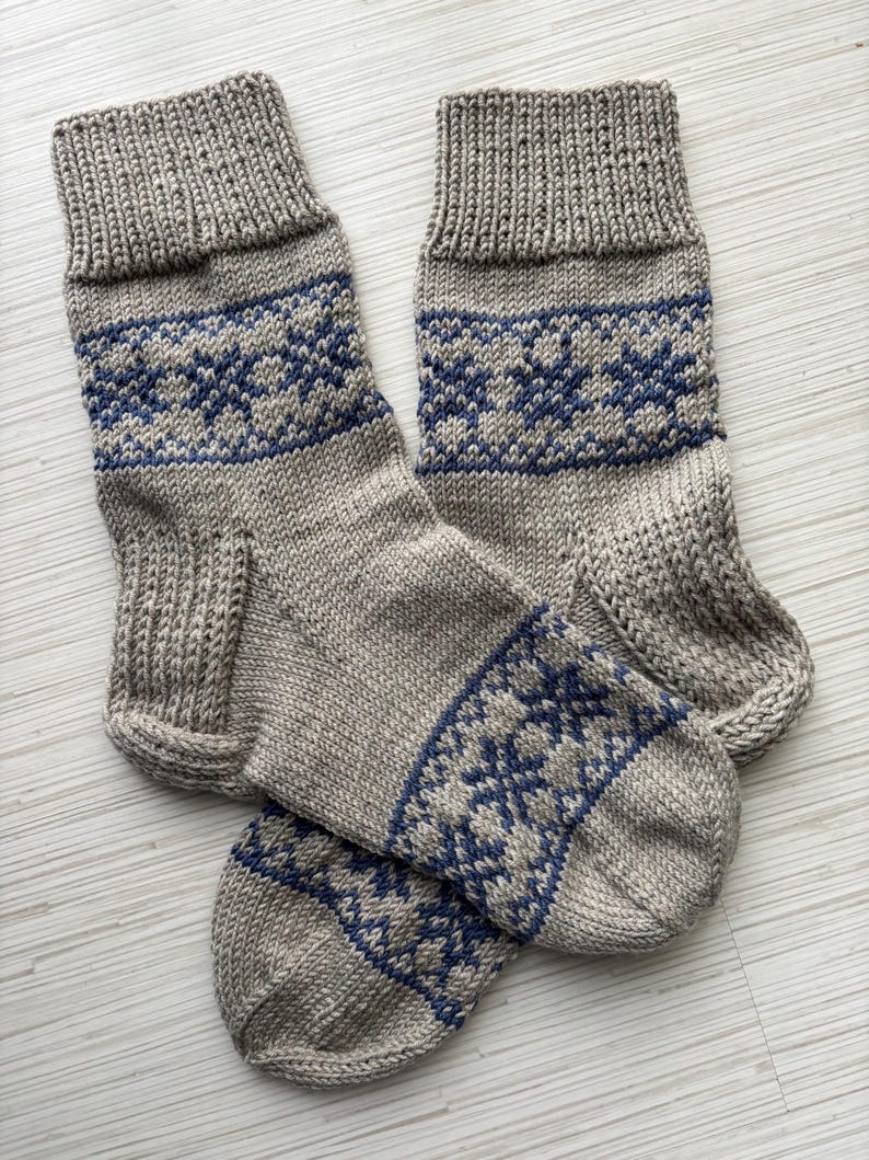 May include: A pair of grey knitted socks with a blue snowflake pattern. The socks are folded together and are lying on a white surface.