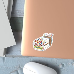 May include: A sticker featuring a cartoon illustration of a loaf of bread with colorful sprinkles and a bow. The text "LET'S GET THIS BREAD" is visible on the sticker. The sticker is placed on a laptop.