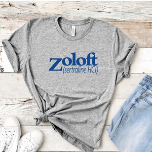May include: A gray t-shirt with the text "Zoloft (sertraline HCl)" printed in blue on the front. The shirt is laid out on a white wooden surface with a pair of white sneakers and blue jeans.