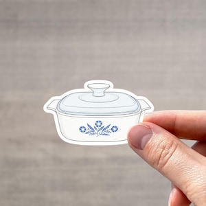 May include: A white casserole dish sticker with a light blue lid and floral design. The sticker is held by a hand against a neutral background. The dish has a classic, vintage aesthetic.