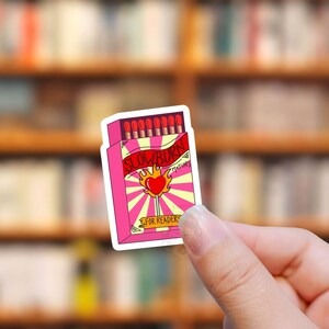 May include: A pink and white matchbox sticker with the text "Slow Burn Matches for Readers". The matchbox has a red heart with flames on a lollipop stick.