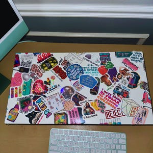 May include: A white rectangular surface covered in colorful stickers with various designs and text. Stickers include phrases like "Please," "High on Life," and "Reject." A computer keyboard is visible at the bottom.