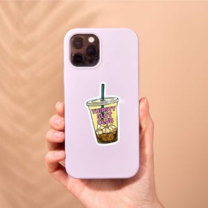 May include: A sticker featuring a cartoon iced coffee drink with the text "Thirsty Slut Club" in a yellow and white font. The drink has brown ice cubes and a green straw. The sticker is on a lavender phone case.