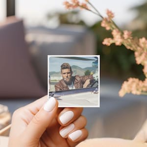 May include: A square photograph featuring a man in a brown leather jacket, sitting in a vintage car. The man is smoking a cigarette. The background shows a mountain range and a blue sky.