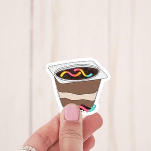 May include: A sticker of a chocolate pudding cup with gummy worms on top. The pudding cup is white with a brown chocolate layer and a white layer on top. The gummy worms are red, yellow, and blue.