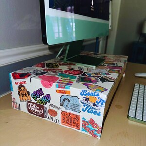 May include: A white box covered in colorful stickers, used as a monitor stand. The stickers feature various designs and text, including "Stay Weird," "Dr. Pepper Slut," and "Boats N' Hoes." A green monitor sits atop the box, with a keyboard visible.