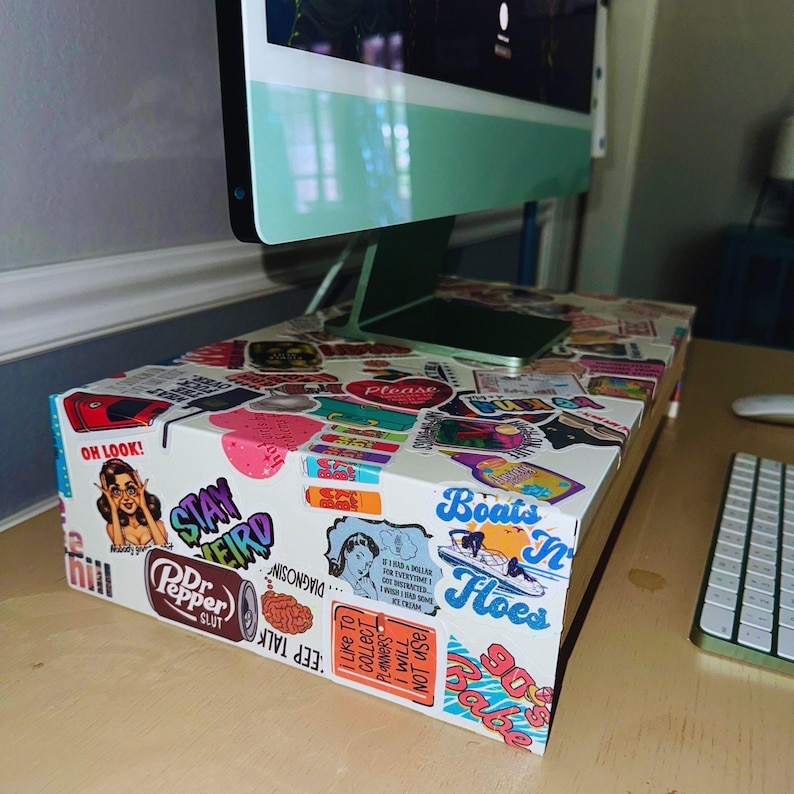 May include: A white box covered in colorful stickers, used as a monitor stand. The stickers feature various designs and text, including "Stay Weird," "Dr. Pepper Slut," and "Boats N' Hoes." A green monitor sits atop the box, with a keyboard visible.