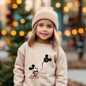 Puede incluir: Una ni&ntilde;a peque&ntilde;a con un su&eacute;ter beige con un gr&aacute;fico de Mickey Mouse negro sosteniendo un globo con el n&uacute;mero 5. El su&eacute;ter tambi&eacute;n tiene el nombre "Evelyn" impreso en &eacute;l. La ni&ntilde;a lleva una boina beige y tiene el pelo rubio.