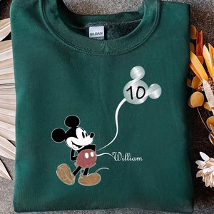 Puede incluir: Sudadera verde con un gr&aacute;fico de Mickey Mouse sosteniendo un globo plateado con el n&uacute;mero 10. El texto "William" est&aacute; impreso debajo del gr&aacute;fico.
