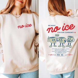 Artistic protest graphic on a sweatshirt featuring retro-style coffee mug characters standing together. The slogan No Ice in our community is prominently displayed in red. The design includes detailed text about brewing justice and community support.