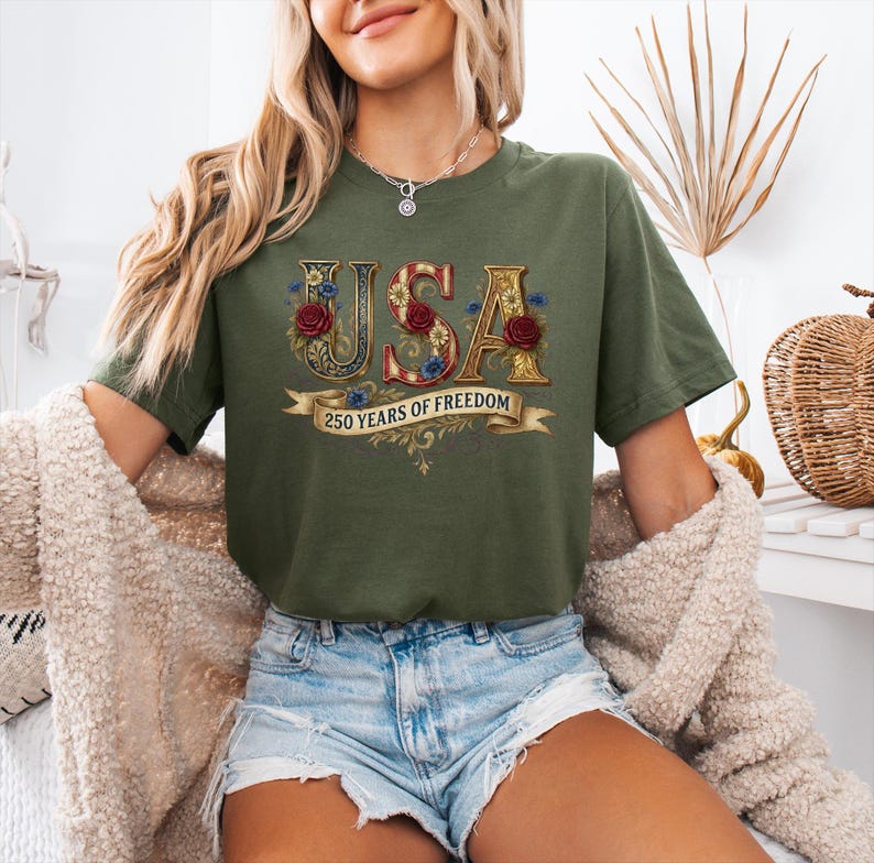 May include: Olive green t-shirt with the word "USA" in a decorative, floral design with red roses and blue flowers. A banner below reads "250 YEARS OF FREEDOM".