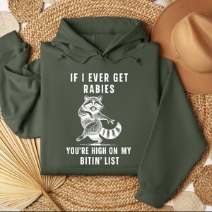Op de afbeelding: Donkergroene hoodie met de tekst "IF I EVER GET RABIES YOU'RE HIGH ON MY BITIN' LIST" en een witte cartoon wasbeer illustratie. De wasbeer heeft een gestreepte staart. De hoodie heeft een capuchon met trekkoord en lange mouwen.