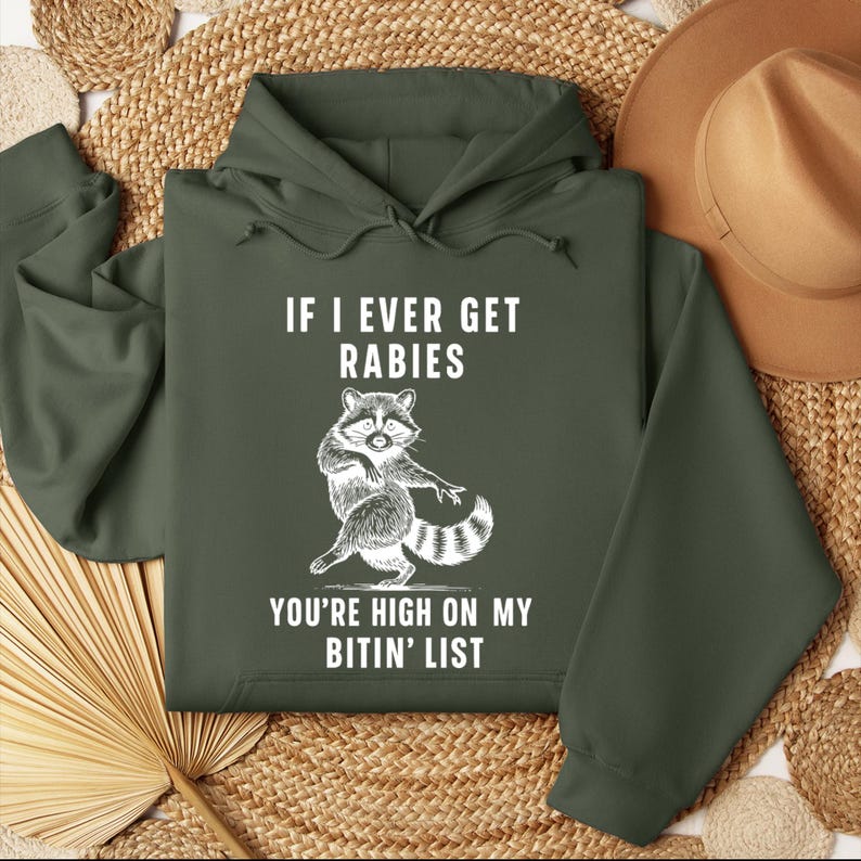Op de afbeelding: Donkergroene hoodie met de tekst "IF I EVER GET RABIES YOU'RE HIGH ON MY BITIN' LIST" en een witte cartoon wasbeer illustratie. De wasbeer heeft een gestreepte staart. De hoodie heeft een capuchon met trekkoord en lange mouwen.