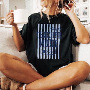 May include: Black t-shirt with a blue and white striped design and the words "TEACHER MODE" in blue. Below the text is an image of an on/off switch with the word "OFF". A white mug is held in the left hand.