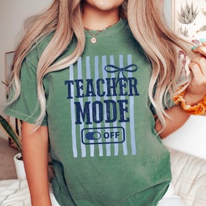 May include: Green t-shirt with the text "TEACHER MODE OFF" in navy blue. The design includes light blue vertical stripes and a bow. The shirt is a casual, comfortable style.