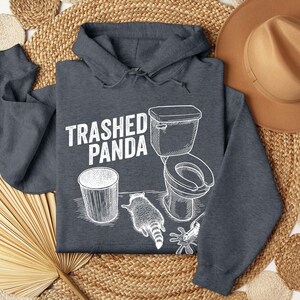 May include: Dark grey hoodie featuring the text "Trashed Panda" and a white graphic of a toilet, bin, and raccoon. The hoodie is made of a soft material and has a hood with drawstrings.