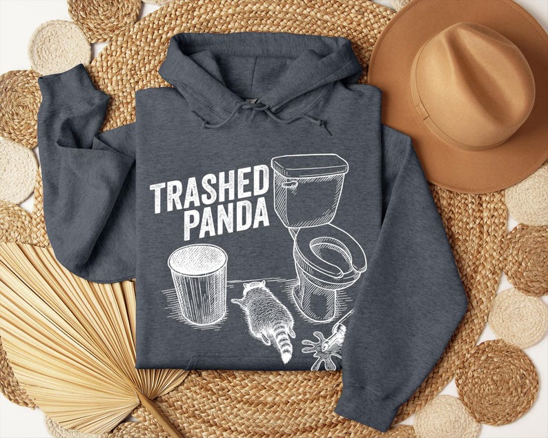 May include: Dark grey hoodie featuring the text "Trashed Panda" and a white graphic of a toilet, bin, and raccoon. The hoodie is made of a soft material and has a hood with drawstrings.
